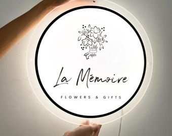 May include: A round, illuminated sign with a white face and black border. The sign features the text "La Mémoire FLOWERS & GIFTS" and a floral illustration. The sign is held by two hands.