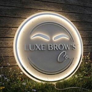 May include: A round, illuminated sign for "Luxe Brow's Co." features a glowing white border and a light gray face. The sign displays stylized eyebrows and the company name in elegant lettering. The sign is mounted on a wooden background, surrounded by grass and flowers.