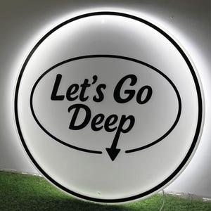 May include: A round, white sign with the text "Let's Go Deep" in black script inside a black oval. A black arrow points downward. The sign has a black border and a glowing white light around the edge.