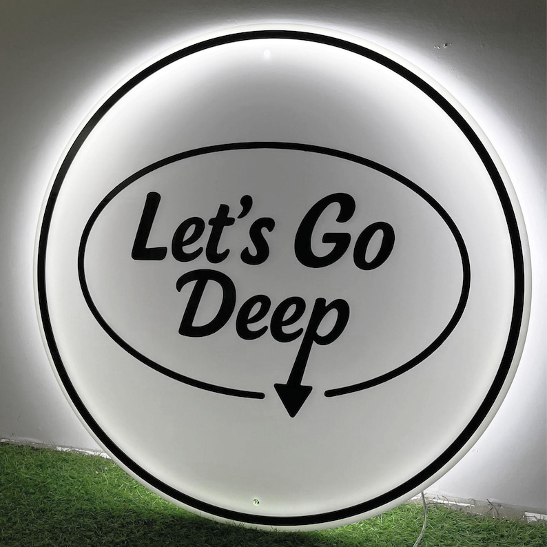 May include: A round, white sign with the text "Let's Go Deep" in black script inside a black oval. A black arrow points downward. The sign has a black border and a glowing white light around the edge.