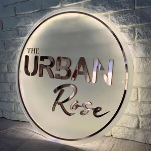 May include: A round, illuminated sign with the text "THE URBAN Rose" in a stylish font. The sign has a rose gold border and lettering, set against a frosted white background. The sign is mounted on a white brick wall.