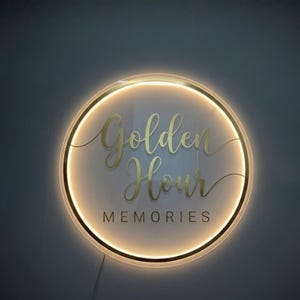 May include: A round, illuminated sign with the words "Golden Hour Memories" in gold script. The sign has a gold border and a warm, glowing light. The background is a dark gray.