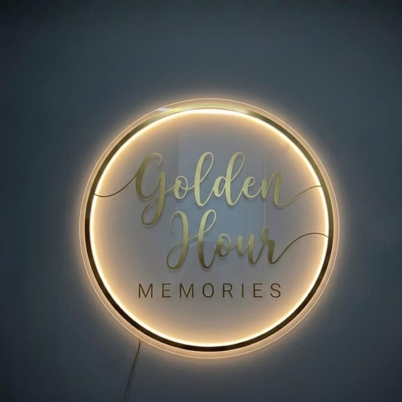 May include: A round, illuminated sign with the words "Golden Hour Memories" in gold script. The sign has a gold border and a warm, glowing light. The background is a dark gray.