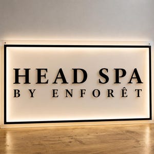 May include: A rectangular wall-mounted sign featuring the black text HEAD SPA BY ENFORÊT in a serif font. The sign has a white background, a black border, and integrated backlighting that creates a soft glow around the perimeter of the display.