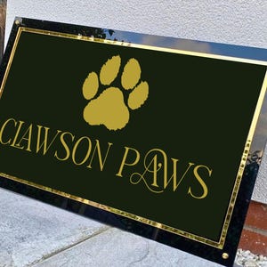 May include: Black rectangular sign with gold trim and the words "CLAWSON PAWS" in gold script. A gold paw print is at the top. The sign is mounted on a wall.