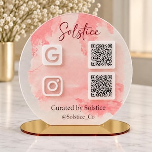 May include: A circular acrylic sign with a pink watercolor wash background, featuring the text Solstice, Curated by Solstice, and @Solstice_Co. It includes white Google and Instagram icons paired with two black and white QR codes on a gold base.