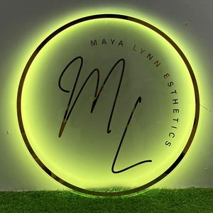 May include: A circular sign with a gold border and a bright green glow. The sign features the text "MAYA LYNN ESTHETICS" in a circular arrangement around a stylized "ML" monogram in black script. The background is a light green.