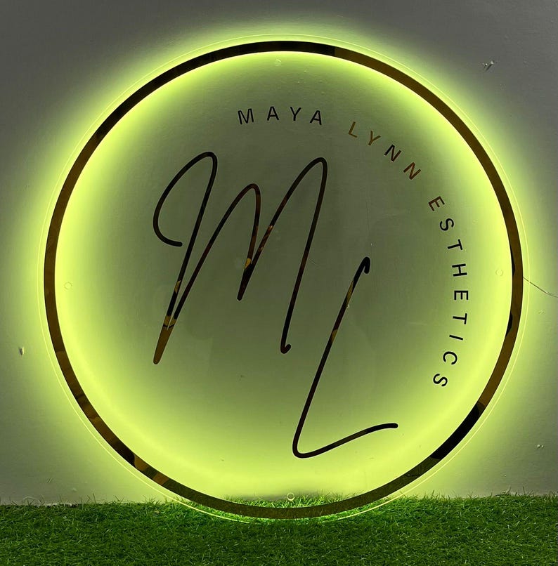 May include: A circular sign with a gold border and a bright green glow. The sign features the text "MAYA LYNN ESTHETICS" in a circular arrangement around a stylized "ML" monogram in black script. The background is a light green.