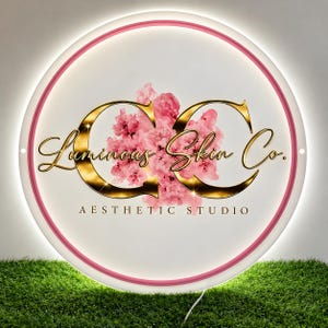 May include: A circular LED sign with a pink border and white background. The center features large gold letters CC, a cluster of pink cherry blossoms, and the text Luminous Skin Co. AESTHETIC STUDIO in black lettering.