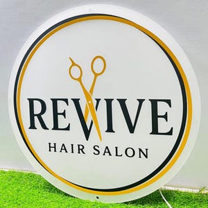 May include: A circular white sign featuring the text REVIVE HAIR SALON in black capital letters. A pair of yellow scissors is positioned vertically behind the letter V. The sign is framed by a thin black and yellow circular border.