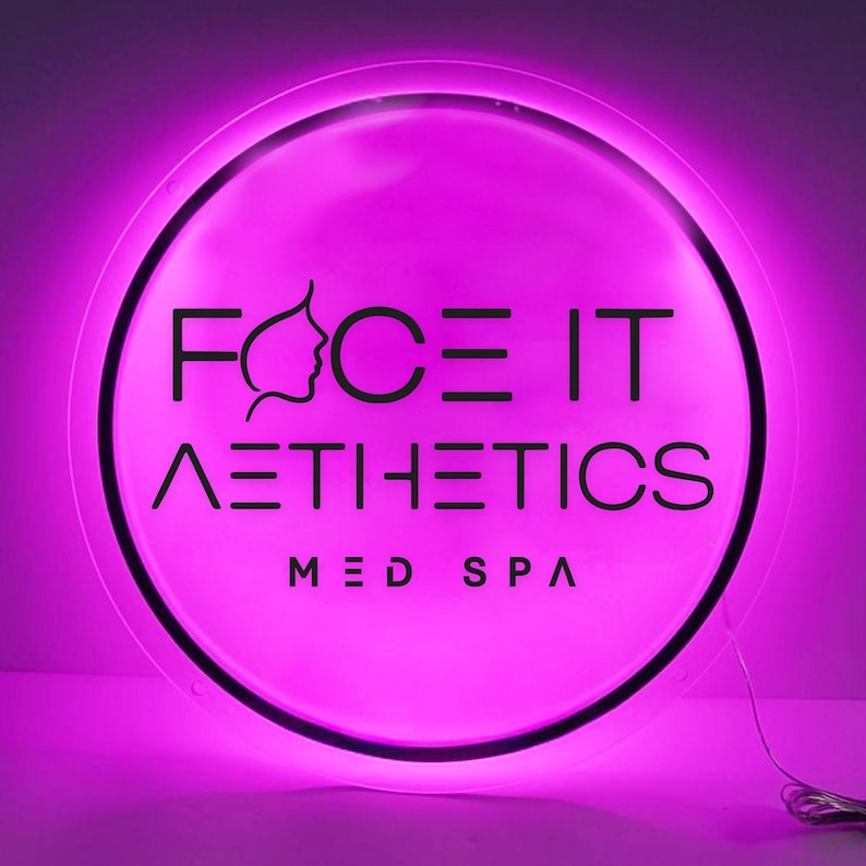 May include: A round, illuminated sign with a bright pink glow. The sign features the words "FACE IT AESTHETICS MED SPA" in black, with a stylized face silhouette. The sign is designed for a medical spa.