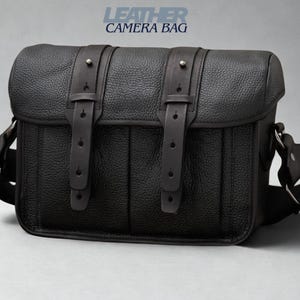 May include: Black leather camera bag with the text "LEATHER CAMERA BAG". The bag has a rectangular shape with a flap closure and two buckle straps. It features a textured surface and a shoulder strap.