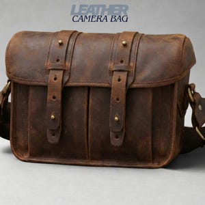 May include: A brown leather camera bag with a flap closure and adjustable shoulder strap. The bag features two front pockets and is secured with two buckle straps. The text "LEATHER CAMERA BAG" is visible.