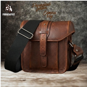 May include: A brown leather camera bag with a black adjustable shoulder strap. The bag has a flap closure with a buckle and a small red accent. The text "Personalised leather Camera bag" is visible.