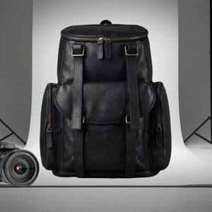 Könnte beinhalten: Schwarzer Lederrucksack mit mehreren Fächern und verstellbaren Trägern. Er verfügt über einen oberen Griff, Reißverschlüsse und goldfarbene Beschläge. Eine DSLR-Kamera mit Objektiv befindet sich im Vordergrund.