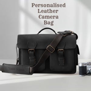 Peut inclure: Sac d'appareil photo en cuir noir avec une bandoulière et deux poches avant. Le sac a une poignée supérieure et des boucles métalliques. Un appareil photo vintage est à côté du sac. Le texte sur le sac dit "Your Name". Le texte au-dessus du sac dit "Personalised Leather Camera Bag".