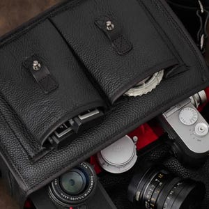 May include: Black leather camera bag with multiple compartments and a red interior. The bag is filled with camera equipment, including lenses and a silver camera body. The bag has a textured surface and a top handle.