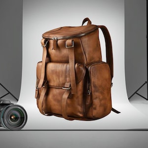 Könnte beinhalten: Ein brauner Lederrucksack mit mehreren Fächern, Riemen und Schnallen. Die Tasche hat einen oberen Griff und gepolsterte Schultergurte. Eine Kamera mit Objektiv befindet sich im Vordergrund. Das Logo "FORGE&FIST" befindet sich in der oberen linken Ecke.