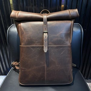 May include: A brown leather backpack with a roll-top closure and a front pocket. The backpack has a buckle closure and adjustable straps. The bag is sitting on a black chair.