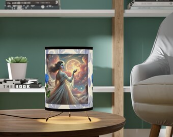 May include: A table lamp with a white fabric shade featuring a painting of a woman in a white dress standing in a field with blue flowers. The lamp has a black base and three legs.