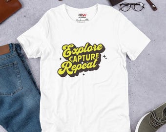 May include: White t-shirt with a retro-style graphic that says "Explore Capture Repeat" in yellow and brown lettering.