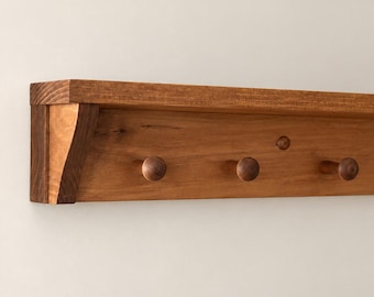 Puede incluir: Un perchero de pared de madera con un estante horizontal y un panel trasero con tres ganchos redondeados. El perchero está fabricado en madera de tono medio con una veta visible e incluye soportes laterales para mayor estabilidad.