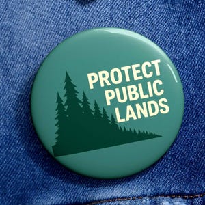May include: A round, teal button with the text "PROTECT PUBLIC LANDS" in white. A dark green illustration of pine trees is on the left. The button is pinned to a blue denim jacket.