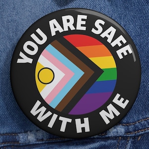 Puede incluir: Un botón negro con el texto blanco "YOU ARE SAFE WITH ME". El botón presenta un diseño de arcoíris y la bandera del Orgullo Progresista. Está prendido a una chaqueta vaquera azul.
