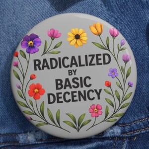 Może przedstawiać: Okrągła, szara przypinka z czarnym napisem "RADICALIZED BY BASIC DECENCY". Przypinka jest ozdobiona kolorowymi kwiatami i zielonymi liśćmi na obrzeżach. Przypinka jest przypięta do dżinsowej kurtki.