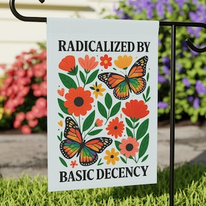 May include: A white garden flag displaying the text "RADICALIZED BY BASIC DECENCY". The flag has a floral design with orange, red, and yellow flowers, green leaves, and two butterflies with rainbow wings.