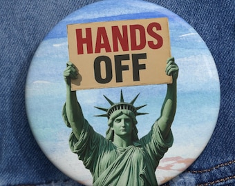 May include: A round button featuring the Statue of Liberty holding a sign that reads "HANDS OFF" in red letters. The button has a blue and white background and is pinned to a denim jacket.