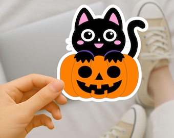 May include: A Halloween sticker showing a black cat sitting on an orange pumpkin with a carved face. The cat has pink ears and a smiling face. The sticker is held in a person's hand. The background includes a laptop and beige trainers.