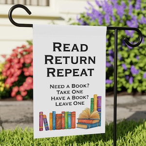 May include: A white garden flag with the text "READ RETURN REPEAT" in black. Below, it reads "NEED A BOOK? TAKE ONE HAVE A BOOK? LEAVE ONE." Colorful book illustrations are at the bottom. The flag is on a black metal stand.