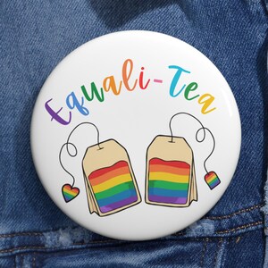 Può includere: Un bottone bianco con la scritta "Equali-Tea" nei colori dell'arcobaleno. Sono raffigurate due bustine di tè con strisce arcobaleno, insieme a dettagli a forma di cuore e quadrato arcobaleno. Il bottone è su uno sfondo di denim.