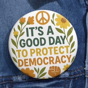 May include: A white button with the text "IT'S A GOOD DAY TO PROTECT DEMOCRACY" in green and orange. The button features a peace sign, flowers, and a fist graphic, set against a denim jacket.