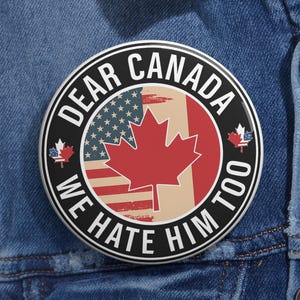 May include: A circular pinback button featuring a black border with the white text "DEAR CANADA WE HATE HIM TOO". The center displays a split design combining the American flag and a red maple leaf on a beige background, with two small maple leaf icons.