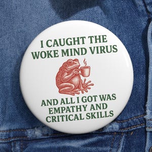 Può includere: Una spilla rotonda bianca con un'illustrazione rossa di una rana che tiene in mano una tazza di caffè. Il testo in verde scuro recita: I CAUGHT THE WOKE MIND VIRUS AND ALL I GOT WAS EMPATHY AND CRITICAL SKILLS. La spilla è fissata su tessuto denim blu.