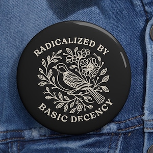 Może przedstawiać: Czarny guzik z białym napisem "RADICALIZED BY BASIC DECENCY" otaczającym białą ilustrację ptaka i kwiatów. Guzik jest umieszczony na dżinsowej kurtce.