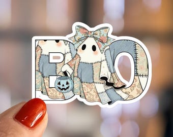 Peut inclure: Autocollant avec le mot « BO » représentant un fantôme et un seau citrouille. Le fantôme porte un costume en patchwork avec un nœud floral. L'autocollant a une bordure blanche.