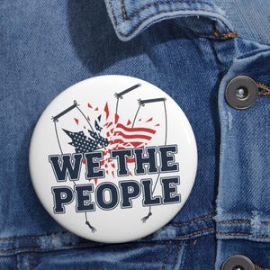 Puede incluir: Un botón blanco con el texto "WE THE PEOPLE" en azul marino, con un gráfico de la bandera estadounidense y un diseño de marioneta en rojo, blanco y azul. El botón está prendido a una chaqueta vaquera.