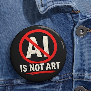 May include: A round pinback button with a black background featuring the white text AI IS NOT ART. A red circle with a diagonal line through it overlays the letters AI. The button is attached to a blue denim fabric with a visible metal button.