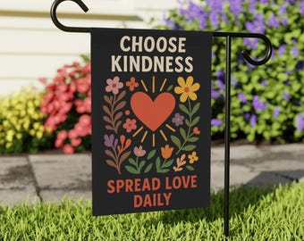 May include: A black garden flag with the words "CHOOSE KINDNESS" and "SPREAD LOVE DAILY" in white and orange text. The flag features a red heart with sun rays, surrounded by colorful flowers and foliage. The flag is displayed on a black metal stand in a garden.