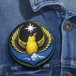 May include: A round button pin featuring a yellow bird with outstretched wings, a white star, and a mountain scene. The design is set against a black and blue background with green and yellow accents. The pin is displayed on a denim jacket.