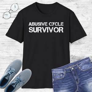 May include: Black t-shirt with the words "ABUSIVE CYCLE SURVIVOR" in distressed white lettering. The shirt is paired with blue jeans and gray shoes. A pair of eyeglasses rests on the surface.