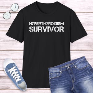 May include: Black t-shirt with the words "HYPERTHYROIDISM SURVIVOR" in white, distressed lettering. The shirt is paired with blue jeans and a pair of blue sneakers. A pair of round eyeglasses rests on a wooden surface.