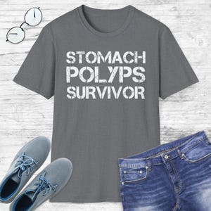 May include: Gray t-shirt with the words "STOMACH POLYPS SURVIVOR" printed in white. The shirt is displayed with blue jeans and gray shoes, and a pair of glasses.