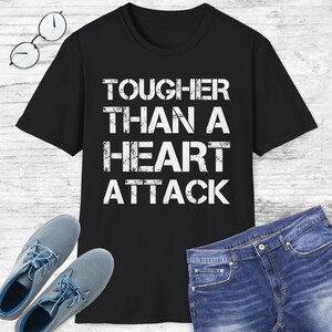 May include: Black t-shirt with the text "TOUGHER THAN A HEART ATTACK" in white, distressed lettering. The shirt is paired with blue jeans and gray shoes. A pair of eyeglasses is also visible.