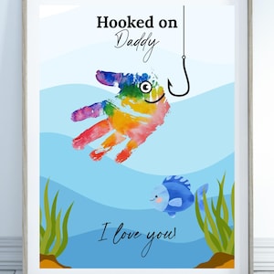 May include: Framed art print featuring a colorful handprint transformed into a fish, hooked by a fishing hook. The text "Hooked on Daddy" and "I love you!" are displayed on a blue and white background with a cartoon fish and aquatic plants.