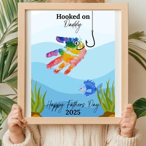 May include: A framed print with the text "Hooked on Daddy" and "Happy Fathers Day! 2025". The artwork features a colorful handprint fish with a fishing hook and a blue fish. The frame is light brown.