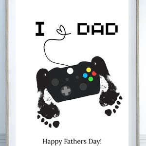 Può includere: Stampa incorniciata con la scritta "I ♥ DAD" sopra un disegno di controller di videogiochi nero con impronte nere. Le parole "Happy Fathers Day!" sono in basso sulla stampa.